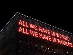 Neon lights on a building that say "All we have is words. All we have is worlds."