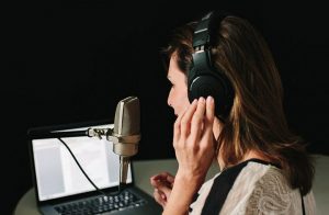 A woman in headphones speaks into a microphone, reading from a laptop