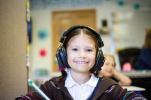 A kid sitting in a classroom wearing bulky headphones grins.