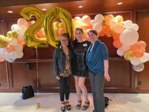 In front of an elaborate helium balloon arc and a gold 20, three women stand smiling at the camera.