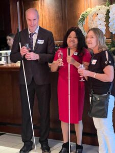 Three people stand side by side, each holding wine glasses. Two people are holding white canes.