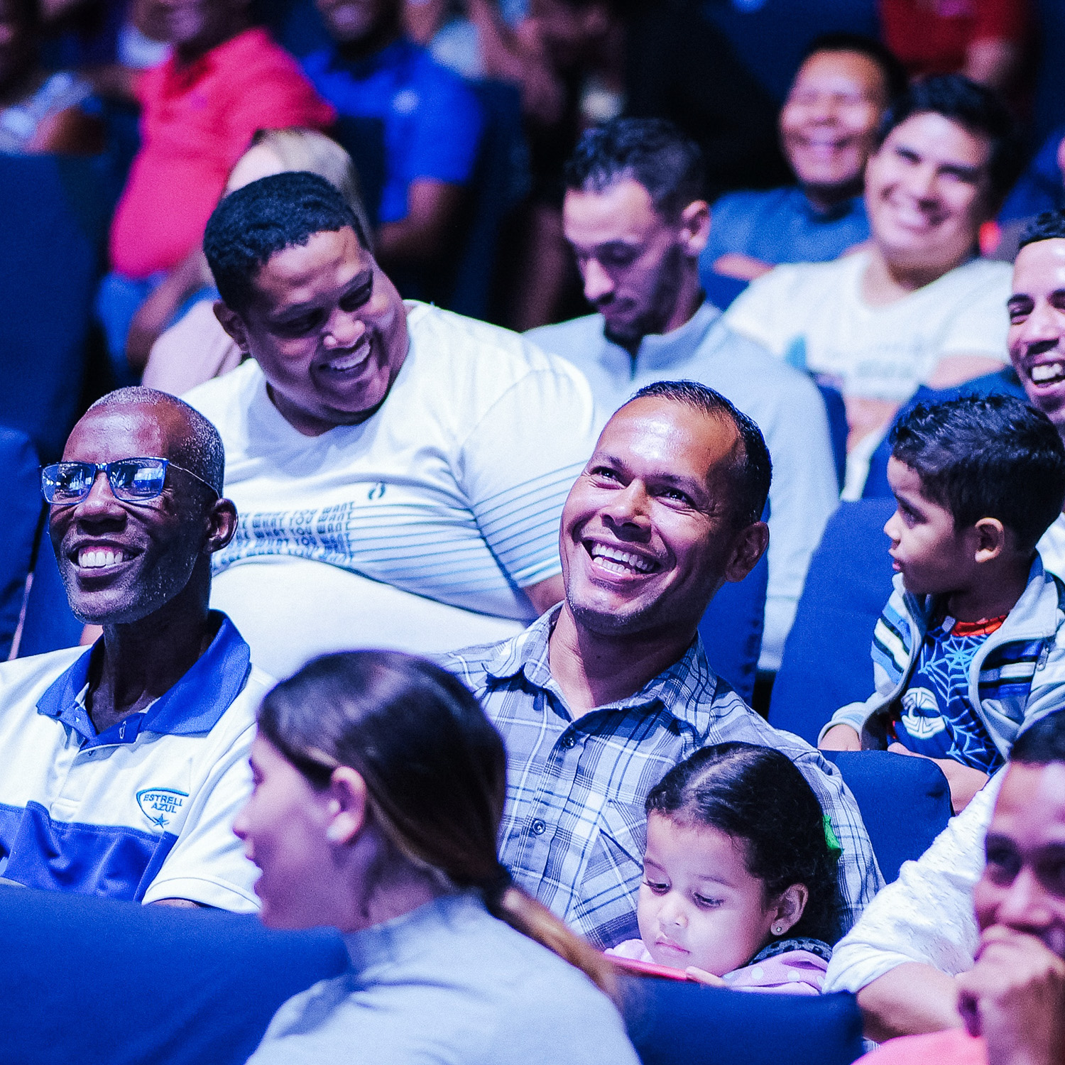 A multiracial crowd of men, women and children sit smiling and laughing in rows of blue upholstered seats.
