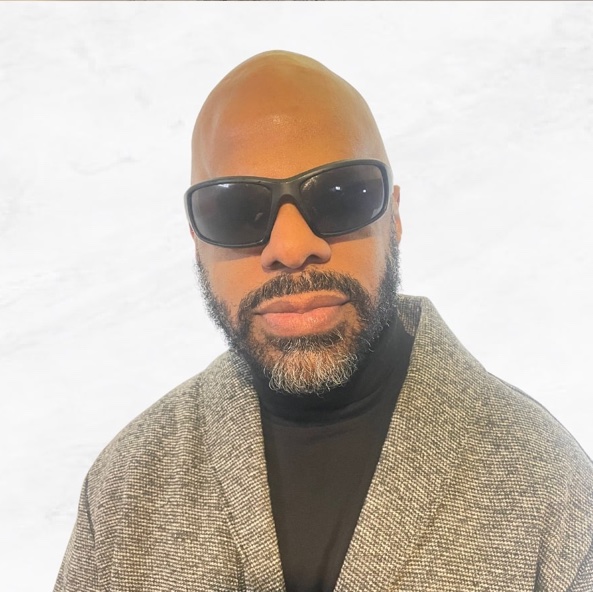 Thomas Reid is a brown skin Black man with a smooth shaven bald head, full neat beard sprinkled with salt and pepper and wearing dark shades.