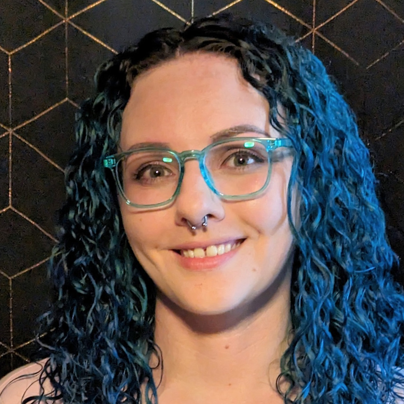 Jenna is a white woman in her early 30s with long, curly blue hair. She wears a pink V-neck t-shirt and square-framed aqua glasses. Her makeup is minimalistic, and a silver septum piercing hangs from her nose.