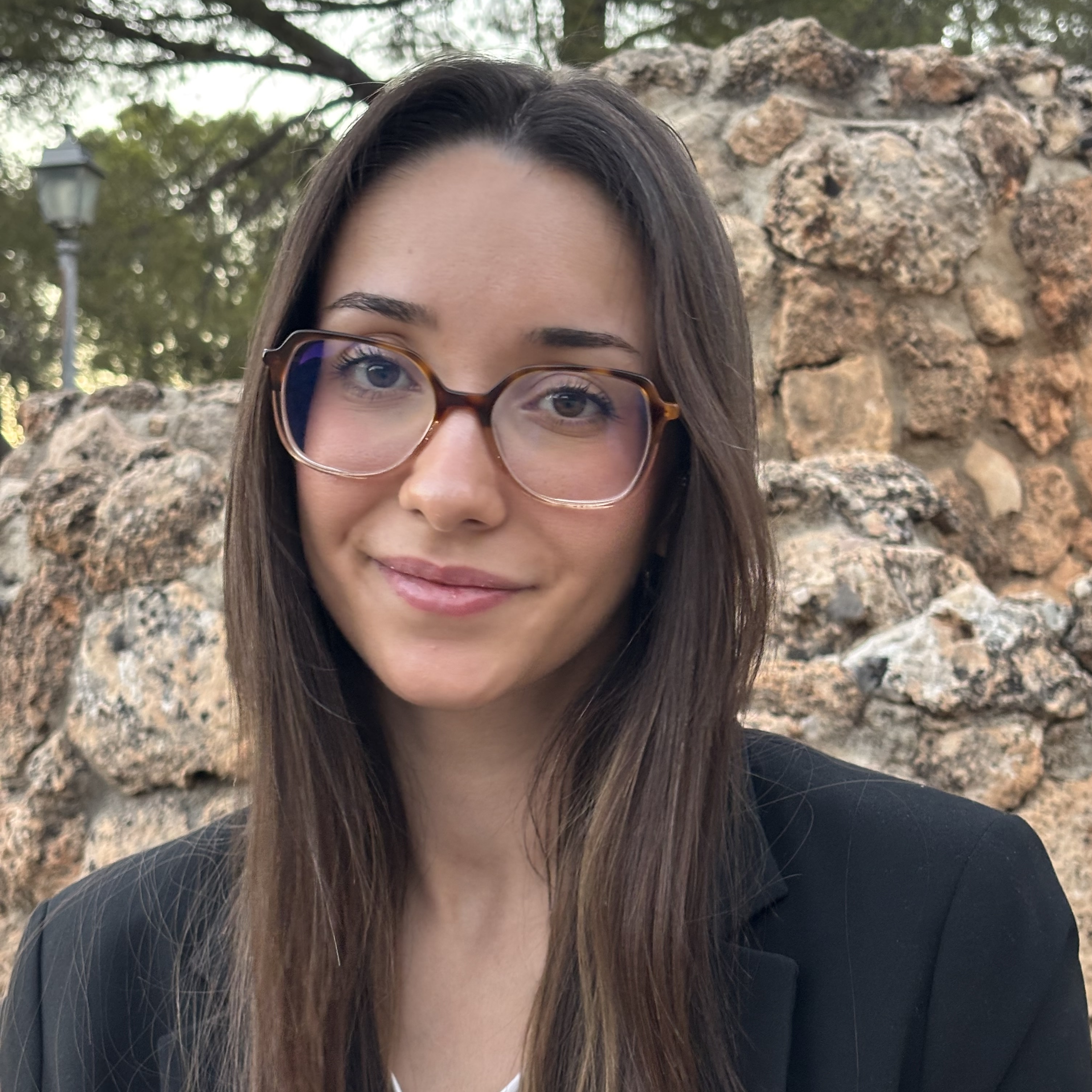 Alicia is a white woman in her mid 20s with long, straight, brown hair. She smiles with full lips, dark eyes and is wearing big brown glasses. She wears light make up, a white t-shirt and a black blazer.